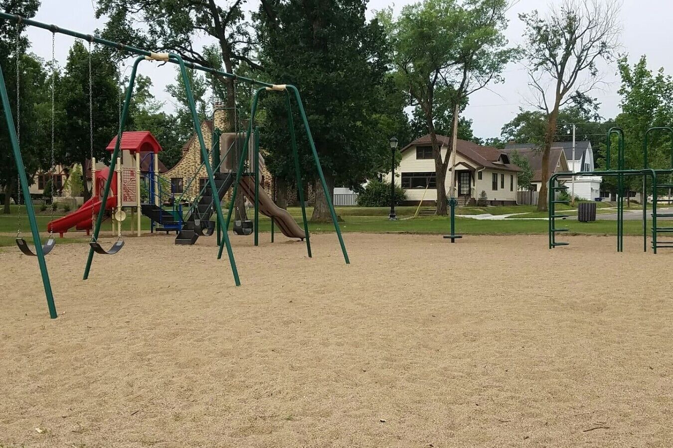 Un parque infantil con columpios y toboganes, rodeado de árboles y casas.