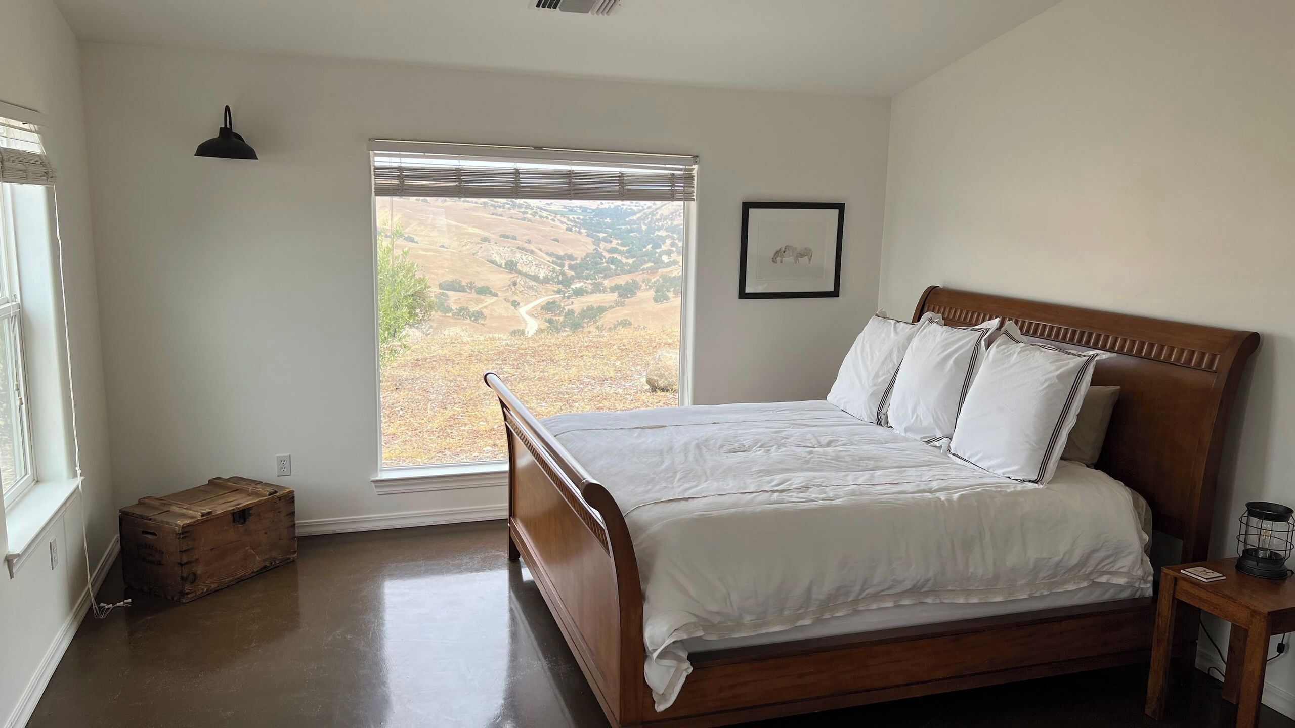A bedroom with a wooden bed, a window with a view of a hilly landscape, a small bedside table, and a wall-mounted light fixture.