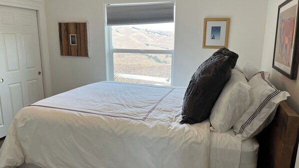 A bedroom with a large bed, a window with a view of hills, a door, and a small picture frame on the wall.