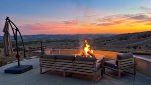An outdoor seating area with a fire, a telescope, and a view of the countryside at sunset.