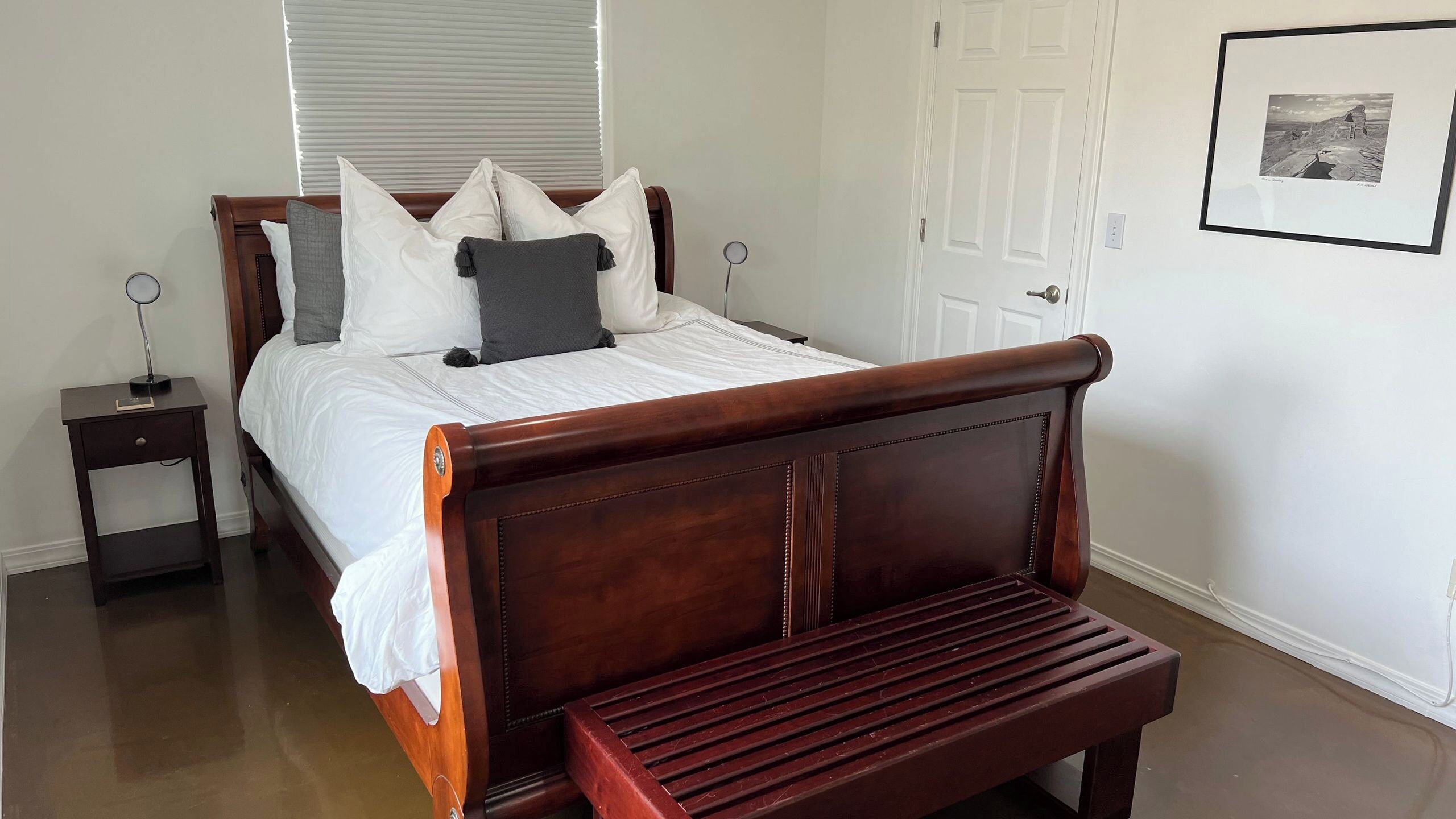 A neatly made bed with white linens and multiple pillows, a wooden bench at the foot of the bed, a bedside table with a lamp, and a framed picture on the wall.