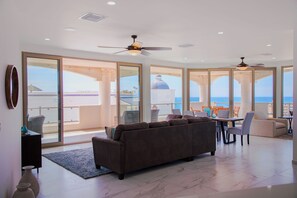 A spacious living room with a large sofa, a dining area with chairs and a table, and a balcony with a view of the sea and a building.