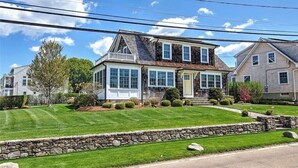 A two-story house with a stone foundation, a front porch, and a well-maintained lawn. - Bonnet Shores Beach Home- 3 Min Walk to Beach! (Narragansett)