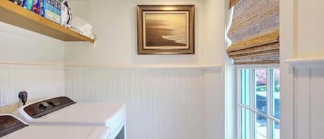 A laundry room with a washing machine, a shelf with cleaning supplies, a framed picture on the wall, and a window with a view.