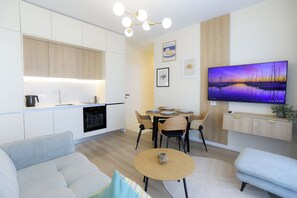 A modern living room with a sofa, a coffee table, and a dining area with chairs and a table. There is a kitchen with white cabinets and a built-in oven. A flat-screen TV is mounted on the wall, and artwork is displayed on the wall. - Seaside Serenity Palm Apartment, Vlore, Albania (Vlore)