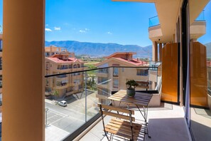 A balcony with a wooden table and chairs, a potted plant, and a view of buildings and mountains.
