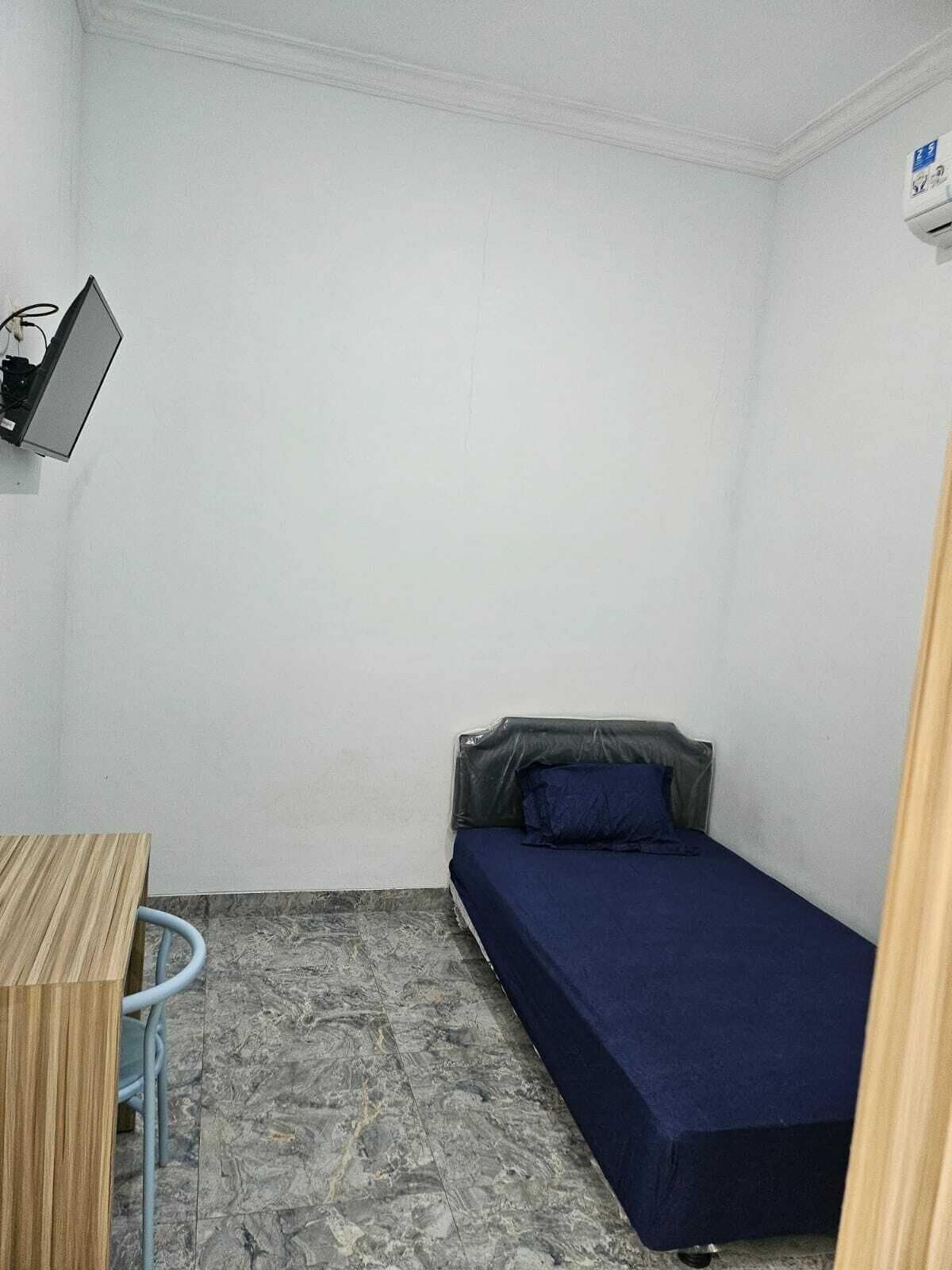 A small, single-bed room with a desk and a chair, a television mounted on the wall, and a tiled floor.