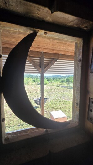 A window with a crescent moon shape, a wooden ceiling, and a view of a grassy field. - Charming 1-bedroom cabin with AC in peaceful Uvalde County on the Nueces River. (Uvalde)