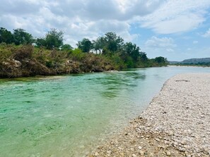 A river with clear water, a pebbly beach, and dense greenery in the background. - Charming 1-bedroom cabin with AC in peaceful Uvalde County on the Nueces River. (Uvalde)