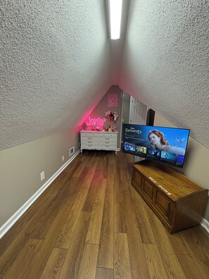 A room with a sloped ceiling, a flat-screen TV on a wooden stand, a white dresser, and a neon sign. - The Perfect Oxford Escape- 4BR Home, 10 Minutes to Football & The Square (Oxford)