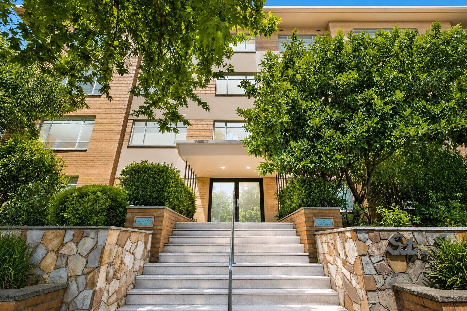 A building with a stone staircase leading to a glass door entrance, surrounded by greenery and a brick facade.