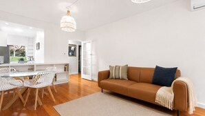 Ein modernes Wohnzimmer mit einem braunen Sofa, weißen Sesseln, einem Glastisch und einer dahinterliegenden Küchenzeile.