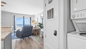 Ein Hotelzimmer mit großem Fenster, einer Sitzecke mit Sessel und Tisch, einer Waschmaschine und einer Tür mit Hinweisschild.