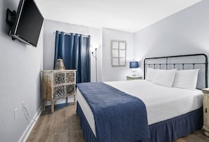 Ein Schlafzimmer mit einem Bett, einem Nachttisch, einer Lampe, einem an der Wand montierten Fernseher und einem Fenster mit Vorhängen.