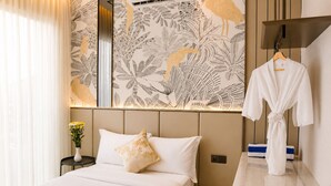 Quarto de hotel com cama, mesa de cabeceira com um vaso de flores, roupão pendurado em um cabide e parede com padrão de folhas.