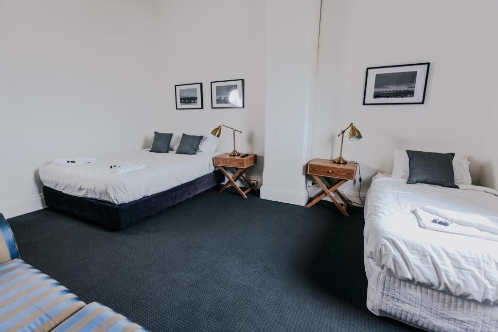 A hotel room with two beds, a small table, a chair, and a wall-mounted lamp.
