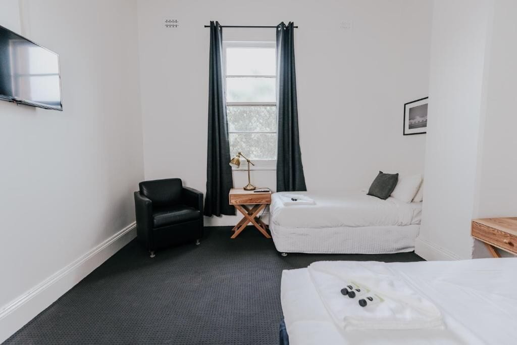 Quarto de hotel com cama, cadeira, mesa pequena, luminária e janela com cortinas.