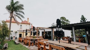 An outdoor dining area with wooden benches and tables, a covered seating area, and a building with a red roof in the background. - Holmesville Hotel (Holmesville)
