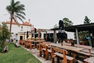 Un área de comedor al aire libre con bancos y mesas de madera, un espacio de estar techado y un edificio con techo rojo al fondo.