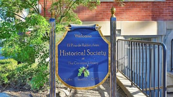 Uma placa azul com letras douradas para a Sociedade Histórica na Biblioteca Carnegie, rodeada por vegetação e um muro de tijolos.