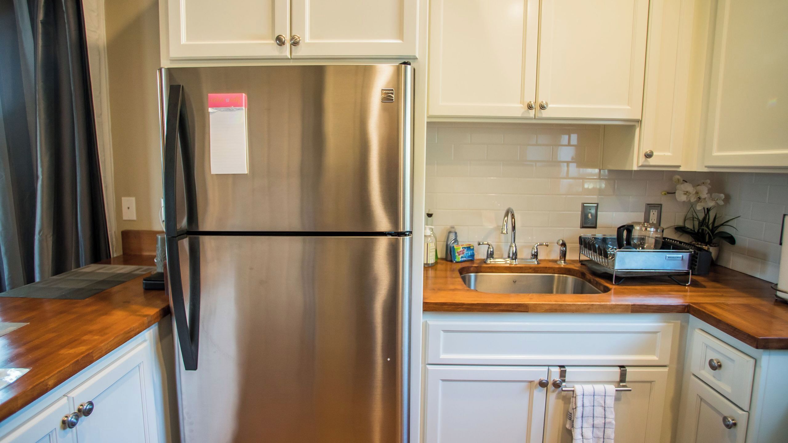 A stainless steel refrigerator in a kitchen with white cabinets, a wooden countertop, and a sink.