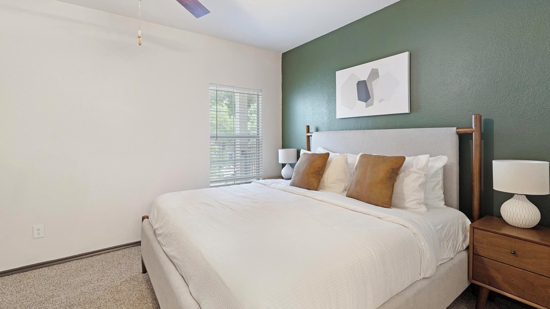 A bedroom with a bed, bedside tables, a window with blinds, and a wall-mounted artwork.