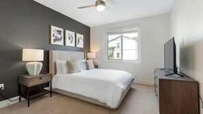 Une chambre à coucher avec un lit, des lampes de chevet, un ventilateur de plafond, un téléviseur posé sur une commode en bois et des œuvres d’art encadrées accrochées au mur.