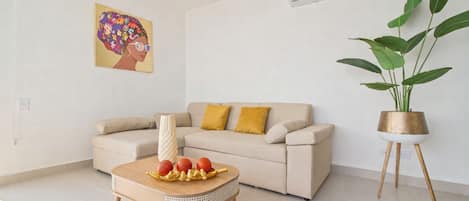 A living room with a beige sofa, a coffee table with decorative items, a potted plant, and a wall-mounted air conditioner.