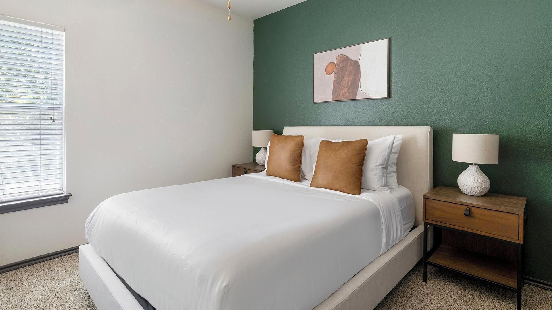 Een net opgemaakt bed met witte lakens, twee nachtkastjes met lampen, een groene accentmuur en een raam met jaloezieën.