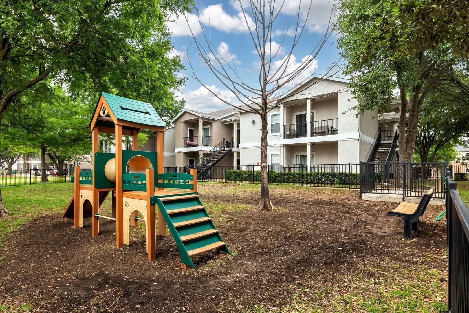 A playground with a slide and climbing structure, surrounded by trees and a building with balconies.