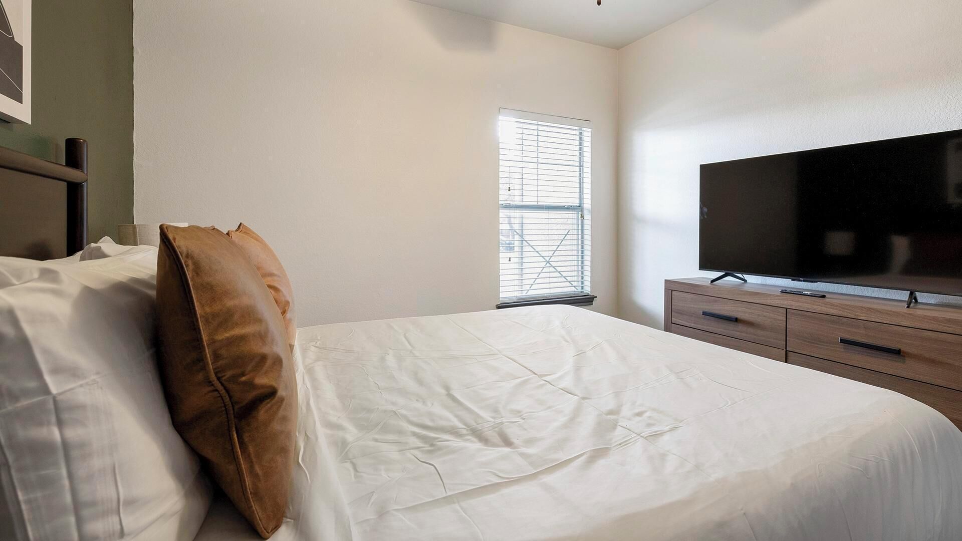 A bedroom with a bed, a television on a wooden dresser, and a window with blinds.