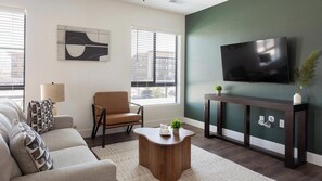 A modern living room with a sofa, armchair, coffee table, and a flat-screen TV mounted on a green wall. - Landing | Incredible 2BD, Game Room, Gym (Goodyear)