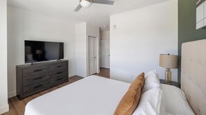 A modern bedroom with a flat-screen TV, a dark wooden dresser, a bed with white linens, a bedside lamp, and a ceiling fan. - Landing | Incredible 1BD, Gym, Courtyard (Goodyear)