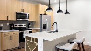 Eine moderne Küche mit Holzausstattung, Edelstahlgeräten, einer großen Kochinsel mit Barhocker und Pendelleuchten.
