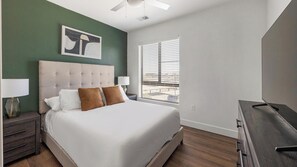 A bedroom with a large bed, two bedside lamps, a ceiling fan, a window with blinds, and a painting on the wall. - Landing | Incredible 2BD, Game Room, Clubhouse (Goodyear)