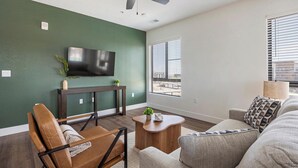 A modern living room with a flat-screen TV mounted on a green wall, a wooden console table, a brown leather chair, a gray sofa, and a small wooden coffee table with a plant. - Landing | Incredible 2BD, Game Room, Clubhouse (Goodyear)