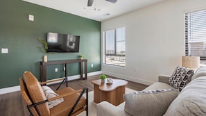 A modern living room with a flat-screen TV mounted on a green wall, a wooden console table, a brown leather chair, a gray sofa, and a small wooden coffee table with a plant. - Landing | Incredible 2BD, Game Room, Clubhouse (Goodyear)