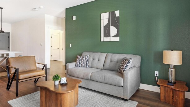 A living room with a grey sofa, a wooden coffee table, a brown leather chair, a green accent wall, and a lamp on a wooden side table. - Landing | Chic 1BD, Clubhouse, Game Room (Goodyear)