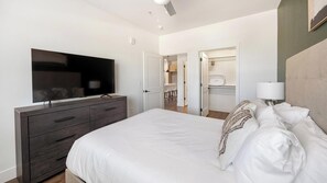 A bedroom with a bed, a TV, a wardrobe, and a bathroom visible through an open door. - Landing | Stylish 1BD, Clubhouse, Game Room (Goodyear)