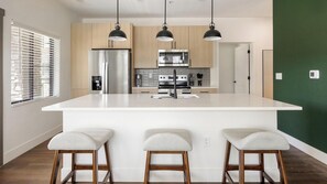 A modern kitchen with a central island, bar stools, and pendant lighting. - Landing | Modern 2BD, Courtyard, Clubhouse (Goodyear)