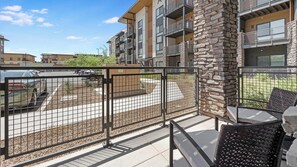 A balcony with a stone pillar, a metal fence, and a view of a parking lot. - Landing | Modern 2BD, Courtyard, Clubhouse (Goodyear)