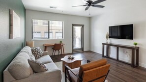 A modern living room with a sofa, a coffee table, a TV, and a ceiling fan. - Landing | Modern 1BD, Clubhouse, Game Room (Goodyear)