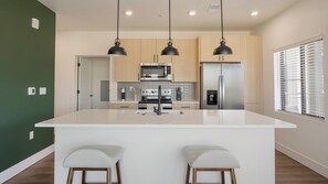 Une cuisine moderne dotée d’un grand îlot central, d’appareils électroménagers en acier inoxydable, de luminaires suspendus et offrant une vue sur l’extérieur grâce à une fenêtre munie de stores.
