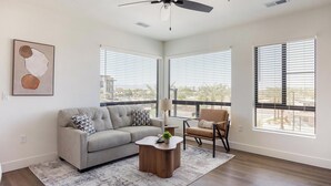 A modern living room with a gray sofa, a wooden coffee table, and a chair. There are large windows with blinds, a ceiling fan, and a framed abstract painting on the wall. - Landing | Stylish 2BD, Gym, Courtyard (Goodyear)
