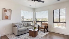 A modern living room with a gray sofa, a wooden coffee table, and a chair. There are large windows with blinds, a ceiling fan, and a framed abstract painting on the wall. - Landing | Stylish 2BD, Gym, Courtyard (Goodyear)