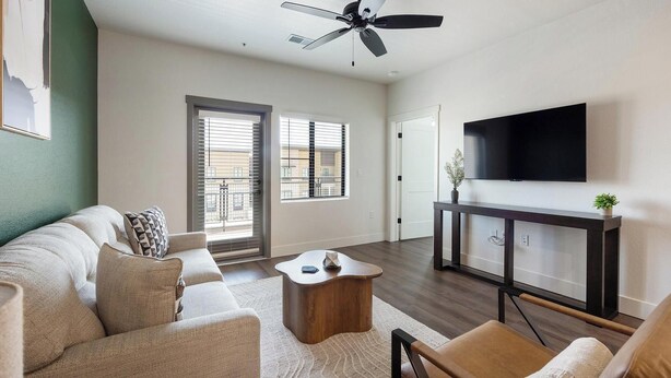 A living room with a sofa, coffee table, and TV stand. There are two windows with blinds, a ceiling fan, and a door. - Landing | Stunning 1BD, Game Room, Gym (Goodyear)