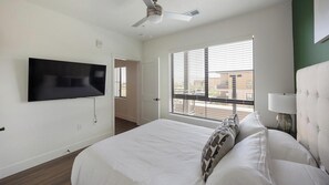 A bedroom with a bed, a television mounted on the wall, a ceiling fan, and a window with blinds. - Landing | Incredible 1BD, Game Room, Gym (Goodyear)
