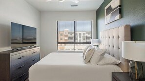 A bedroom with a bed, a TV, a dresser, a window with blinds, and a wall-mounted lamp. - Landing | Stylish 2BD, Gym, Game Room (Goodyear)