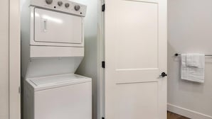 A compact laundry room with a white washing machine and dryer, a closed door, and a towel hanging on a hook. - Landing | Modern 2BD, Game Room, Gym (Goodyear)