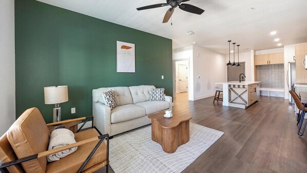 A modern living room with a sofa, coffee table, and a ceiling fan. - Landing | Stylish 2BD, Gym, Game Room (Goodyear)